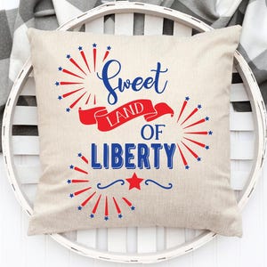 May include: A beige square pillow with the text "Sweet Land of Liberty" in blue script and block letters, with red and blue starburst and ribbon accents. The pillow is on a white, round tray.