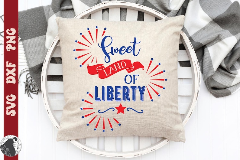 May include: A beige square pillow with the text "Sweet Land of Liberty" in blue script and block letters, with red and blue starburst and ribbon accents. The pillow is on a white, round tray.