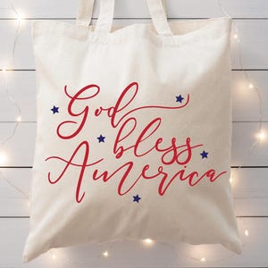 May include: A cream-colored tote bag with the words "God bless America" in red script and blue star accents. The bag is made of a sturdy fabric and has two handles.