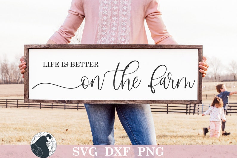 Op de afbeelding: Een wit houten bord met de tekst "Life is better on the farm" in zwarte cursieve letters. Het bord wordt vastgehouden door een persoon die een roze shirt en een blauwe spijkerbroek draagt. Het bord staat voor een groen veld met een houten hek.