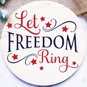 May include: A round wooden sign with the words "Let Freedom Ring" in red and blue script. Red stars and decorative swirls accent the text. The sign has a light wood grain background.
