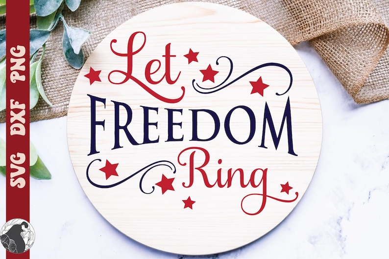 May include: A round wooden sign with the words "Let Freedom Ring" in red and blue script. Red stars and decorative swirls accent the text. The sign has a light wood grain background.