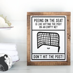May include: A wooden sign with a black and white illustration of a hockey net and puck. The sign reads "Peeing on the seat is like hitting the post on an empty net. Don't hit the post!"