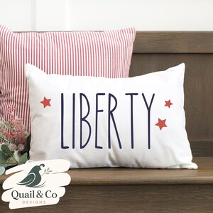 May include: White rectangular pillow with the word "LIBERTY" in navy blue, flanked by three red stars. A second pillow with red and white stripes is behind it. The pillows are on a wooden surface.