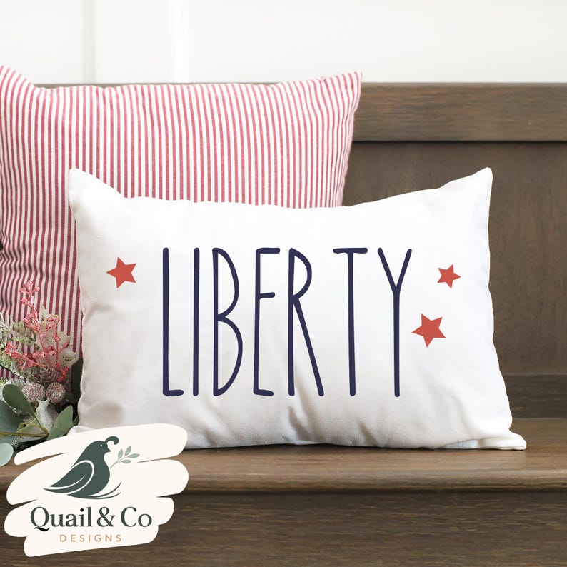May include: White rectangular pillow with the word "LIBERTY" in navy blue, flanked by three red stars. A second pillow with red and white stripes is behind it. The pillows are on a wooden surface.