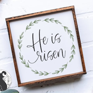 Pu&ograve; includere: Un cartello di legno bianco con cornice marrone presenta una ghirlanda di foglie verdi con il testo "He is risen" in lettere corsive nere.