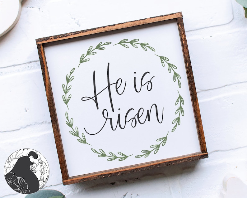 Pu&ograve; includere: Un cartello di legno bianco con cornice marrone presenta una ghirlanda di foglie verdi con il testo "He is risen" in lettere corsive nere.