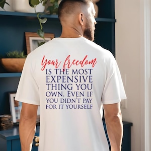 May include: A white t-shirt with the text "Your freedom is the most expensive thing you own. Even if you didn't pay for it yourself" in red and blue. The shirt is worn by a person.