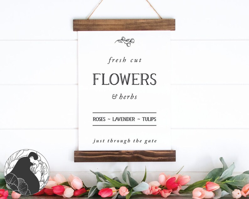 May include: A white sign with black text that reads "fresh cut FLOWERS & herbs ROSES - LAVENDER - TULIPS just through the gate". The sign is hanging from a wooden frame and is against a white wood background.