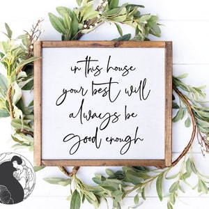 以下が含まれることがあります： 黒い筆記体で「In this house your best will always be good enough」と書かれた白い木製の看板。看板は緑の葉のガーランドで囲まれています。