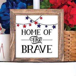 May include: A wooden framed sign with the text "HOME OF The BRAVE" in black. The sign is decorated with red and blue star lights. The background includes a basket of flowers, an American flag, and a "USA" mug.