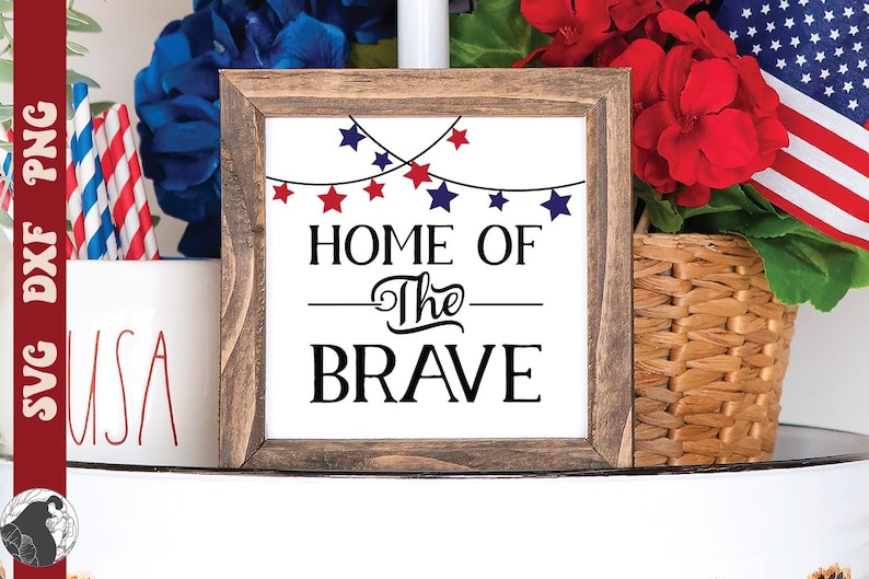 May include: A wooden framed sign with the text "HOME OF The BRAVE" in black. The sign is decorated with red and blue star lights. The background includes a basket of flowers, an American flag, and a "USA" mug.