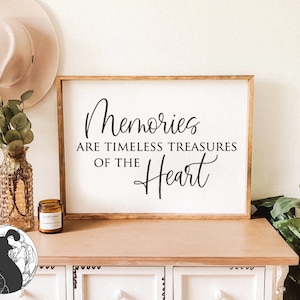 Może przedstawiać: Drewniany znak z tekstem "Memories are timeless treasures of the heart" w czarnym, ozdobnym pismie. Znak stoi na białej komodzie z wazą z zielenią, kapeluszem i świecą.