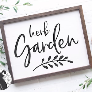 Op de afbeelding: Een wit houten bord met een bruin frame, met de tekst "herb garden" in zwarte cursieve letters met een decoratieve bladeren tak eronder.