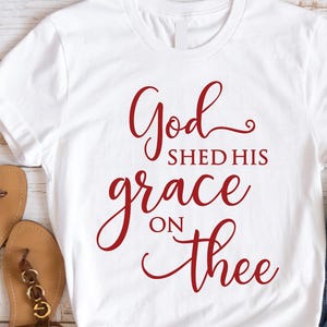 May include: White t-shirt with the red text "God shed his grace on thee." The shirt is paired with brown sandals, blue jeans, and a small American flag. The design is suitable for patriotic or religious themes.