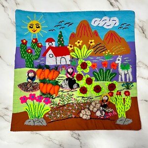 May include: A colorful embroidered pillow featuring a whimsical scene of a village with houses, mountains, flowers, and people. The pillow is made with a variety of fabrics and textures, including felt, cotton, and embroidery thread.
