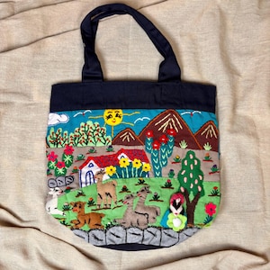 May include: Embroidered tote bag with a navy blue top and handles. The bag features a colourful landscape scene with mountains, a sun, and various animals. The design includes a house, trees, and flowers, creating a vibrant and detailed image.