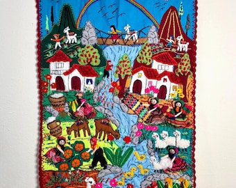 May include: Embroidered wall hanging showing a colourful village scene. The artwork features a blue sky with clouds and a rainbow, red-roofed houses, figures, animals, and lush greenery. The border is a deep red with a scalloped edge.