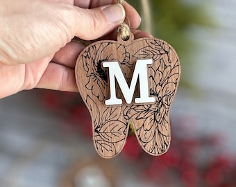 May include: Wooden tooth-shaped ornament with floral engraving and a white letter "M". The ornament is held by a string and features a natural wood grain finish. The floral design is etched in black.