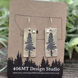 May include: A pair of rectangular brass earrings with a tree design. Each earring has a dark tree silhouette and a small circle at the top. The earrings are displayed on a brown card with a forest silhouette and the text "406MT Design Studio".