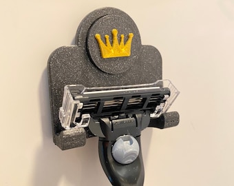 May include: A black 3D printed razor holder with a gold crown design. The holder has a clear plastic razor holder and a black hook for hanging.