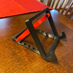 May include: A black and red 3D-printed phone stand. The stand has a rectangular base with multiple grooves, two angled supports, and a red adjustable arm. The stand is designed to hold a phone or tablet.