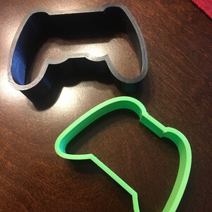 May include: Two plastic cookie cutters shaped like video game controllers. The top cutter is black and the bottom cutter is green.