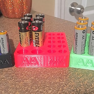 May include: Three 3D printed battery organizers, one black, one red, and one green, each with the word "AAA" printed on the front. The organizers are filled with AA batteries, including brands like "Energizer", "Eveready", and "AmazonBasics".