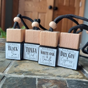 May include: Four small wooden car air fresheners with black labels and black string hangers. The labels read "Rustic Roost Co." and list different scents: "Black Sea", "Tonka & Oud", "White Oak & Vanilla", and "Dry Gin & Cypress".