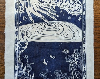 May include: A blue and white woodcut print of a hand reaching down into a body of water. The hand is creating ripples in the water. The water is surrounded by a border of stylized plants and sea creatures. The text "Excuse me, I have work to do" is at the bottom of the print.