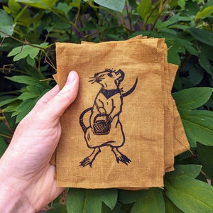 May include: A stack of four brown linen fabric squares with a black and white illustration of a rat holding a basket. The rat is wearing a bandana.