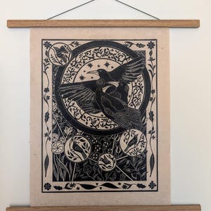 May include: A black and beige linocut print featuring three crows in flight within a circular frame, surrounded by smaller circular bird motifs and floral patterns. The artwork is mounted between two horizontal wooden bars with a hanging cord.