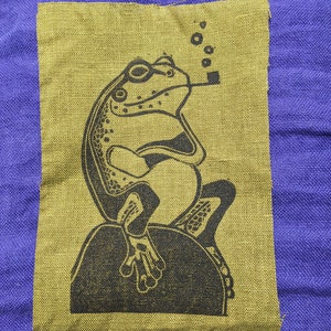 May include: A green fabric patch with a black and white illustration of a frog smoking a pipe. The frog is sitting on a round object. There are small white circles above the frog's head.