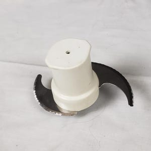 Może przedstawiać: Ostrze robota kuchennego z białą plastikową podstawą i dwoma zakrzywionymi ostrzami ze stali nierdzewnej. Ostrza przeznaczone są do siekania i mieszania składników spożywczych. Przedmiot umieszczony jest na białym tle.