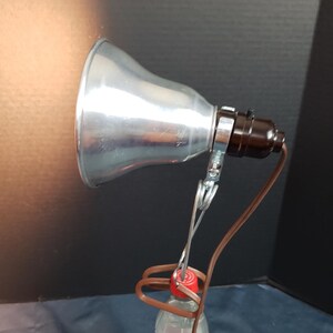 Könnte beinhalten: Ein silberner Lampenschirm aus Metall mit einem schwarzen Stromkabel und einer Klemme, die an einer durchsichtigen Plastikwasserflasche befestigt ist.