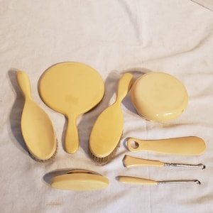 May include: A vintage vanity set in a cream color, including a hand mirror, two brushes, a powder compact, a small dish, and two manicure tools. The set is arranged on a white surface.
