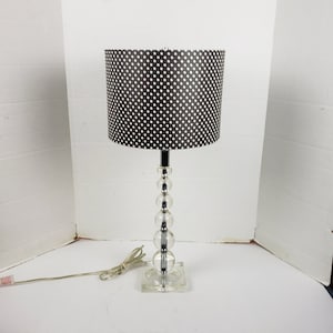 Op de afbeelding: Een tafellamp met een heldere glazen voet en een zwart-witte lampenkap met stippen.