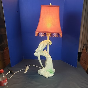 May include: A vintage table lamp featuring a white ceramic parrot perched on a branch. The lamp has a coral-colored lampshade with beaded fringe. The base is white with green accents. The lamp is plugged in and ready to use.