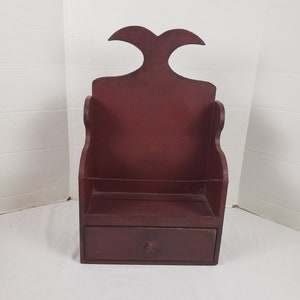 May include: A small, red wooden shelf with a single drawer and a unique, curved top. The shelf is painted a deep red color and has a rustic, antique look.
