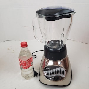 May include: A silver Oster blender with a glass jug and black lid. The blender has multiple speed settings and a clear glass jug. A clear plastic bottle with a red cap is next to the blender.