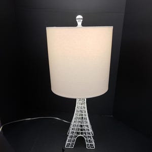 May include: White table lamp with a white fabric lampshade and a base shaped like the Eiffel Tower. The base is made of metal and has a crystal-like finish.