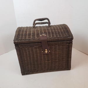 May include: A brown wicker picnic basket with a curved top and a handle. The basket has a leather strap closure with a gold-colored clasp. The woven texture and dark brown color give it a vintage look.