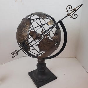 May include: A decorative metal globe with a dark bronze finish. The globe features a wireframe design with continents and is mounted on a black base. An arrow with decorative detailing points through the globe, adding a unique touch.