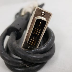以下が含まれることがあります： 銀色のコネクタと黒いケーブルを備えたDVIケーブルのクローズアップ。コネクタには金色のピンとネジが付いています。ケーブルはコイル状で、テキストが印刷されています。