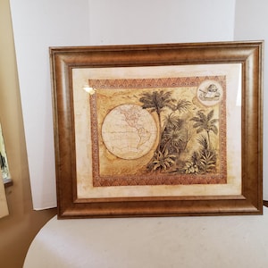 May include: Framed wall art featuring a vintage-style map of the world with a focus on the Americas. The artwork includes illustrations of palm trees and a ship, set within a decorative border. The frame is a warm brown color.