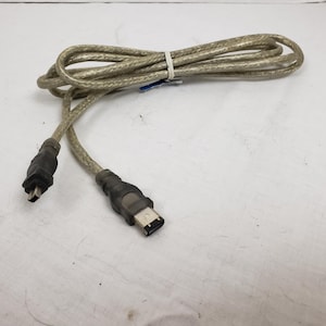 Puede incluir: Un cable FireWire beige y negro, enrollado y asegurado con una brida blanca. El cable tiene dos conectores negros en cada extremo. El cable está sobre una superficie blanca.