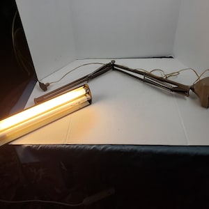 Könnte beinhalten: Eine Gelenk-Schreibtischlampe mit rechteckiger Leuchte und zwei Leuchtstoffröhren. Die Lampe hat eine braune Metalloberfläche und einen Klemmfuß. Die Lampe ist ausgefahren und auf einer weißen Oberfläche positioniert.