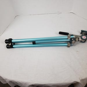 Puede incluir: Un trípode azul claro plegado con herrajes negros y plateados. El trípode tiene tres patas y un asa negra. El trípode está diseñado para fotografía y videografía.