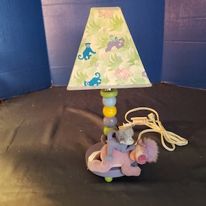 Puede incluir: Una lámpara para niños con una pantalla de lámpara de temática de jungla verde y blanca. La base es de madera y tiene un esquema de color morado, azul, amarillo y verde. Un elefante de peluche morado con un mono azul en la espalda se sienta en la base.