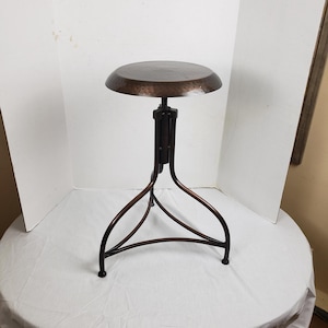 May include: A copper-colored metal adjustable height stool. The round seat has a hammered texture. The base has three curved legs and a central support column. The stool is designed for stability and style.
